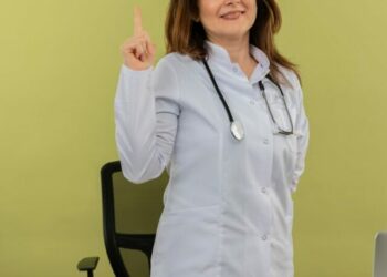 smiling-middle-aged-female-doctor-wearing-medical-robe-stethoscope-standing-desk-with-medical-tools-laptop-raising-finger-keeping-hand-back-isolated_141793-69177