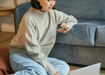 vertical-shot-of-young-woman-in-cozy-home-working-on-laptop-using-smartphone-and-drinking-coffee-sitting-on-floor-near-sofa-free-photo