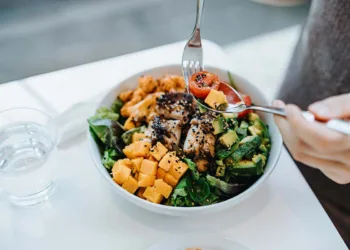 Close up, high angle shot of young woman enjoying multi-coloured healthy fruit, vegetables with grilled chicken salad bowl with balanced nutrition in cafe, with a glass of water by the side. Healthy eating lifestyle. People, food and lifestyle concept