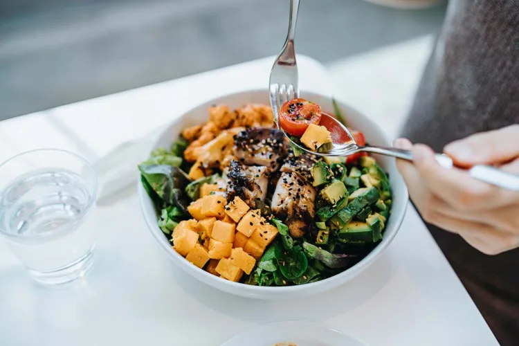 Close up, high angle shot of young woman enjoying multi-coloured healthy fruit, vegetables with grilled chicken salad bowl with balanced nutrition in cafe, with a glass of water by the side. Healthy eating lifestyle. People, food and lifestyle concept