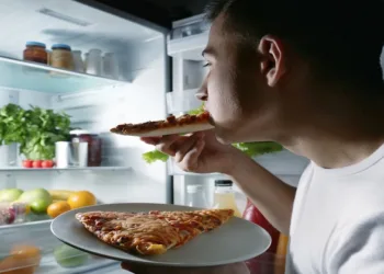 Young man eating pizza near refrigerator at night