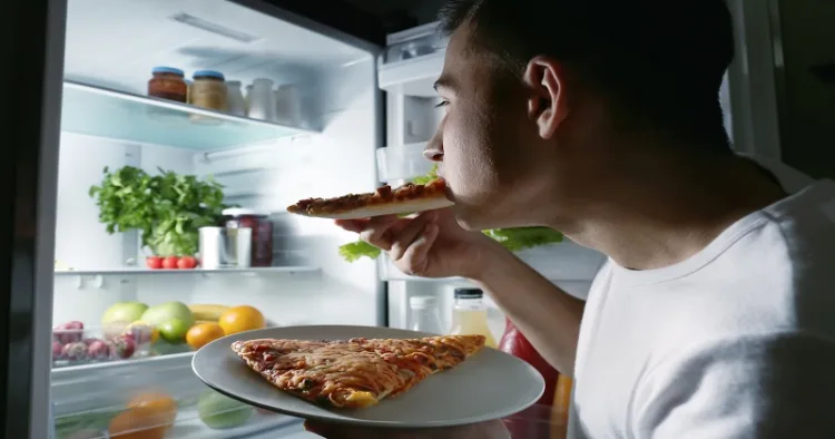 Young man eating pizza near refrigerator at night